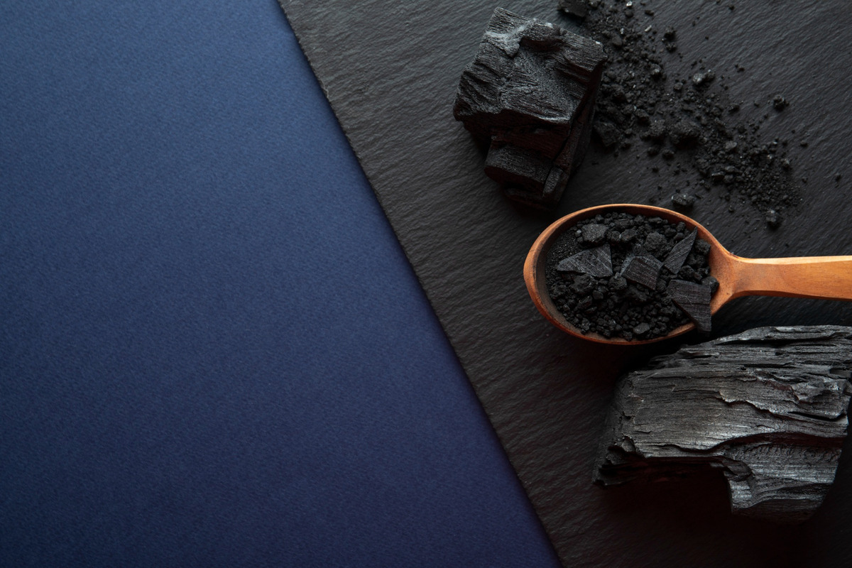Flat lay image featuring pieces of black charcoal and a wooden spoon filled with powdered charcoal on a dark slate surface. The background includes a contrasting dark blue texture, creating a moody, earthy aesthetic. Highlights charcoal’s association with